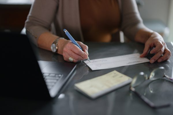 A close-up of a woman writing a personal check at a desk, accompanied by a laptop, reading glasses, and other items, representing finance, payment, and attention to transactions.